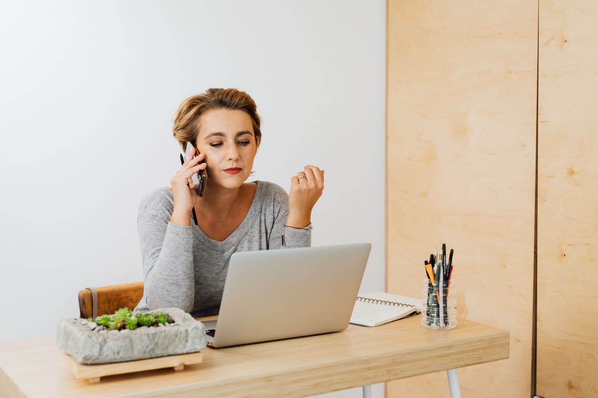 A person working from a home office desk on a laptop while taking a phone call, representing a home-based travel agent business